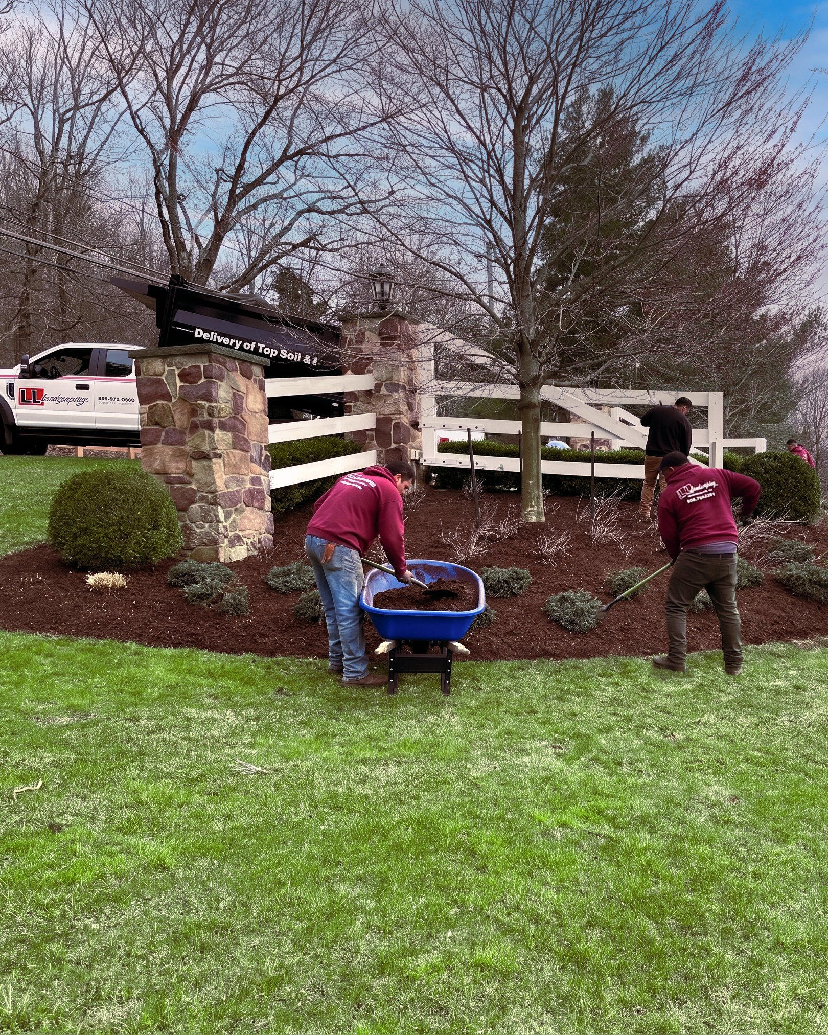 Two LL Landscaping crew members in red jackets working a curved mulch bed — one bending over a blue wheelbarrow, one raking — along a white split-rail fence, with the LL work truck and a stone-pillar gateway in the upper left.