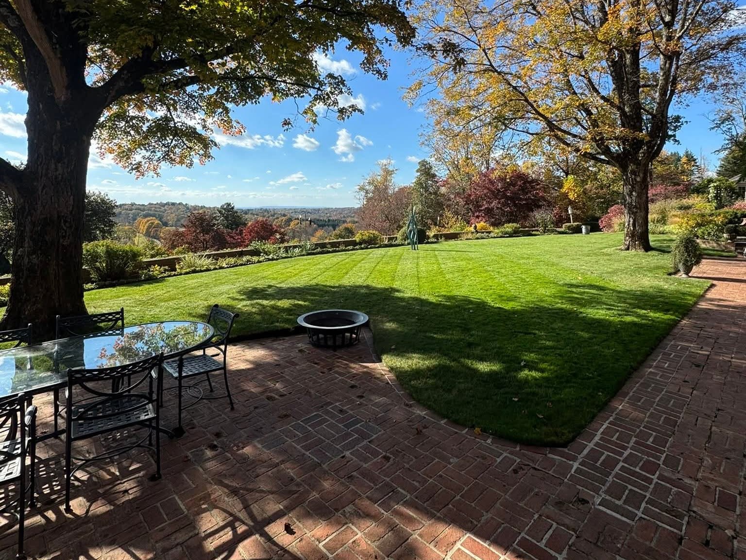An estate-style maintained lawn at golden hour with a small stone fire pit centered on tightly cut grass, a brick patio with iron furniture in the foreground, and mature autumn-color trees framing both sides above a distant valley.