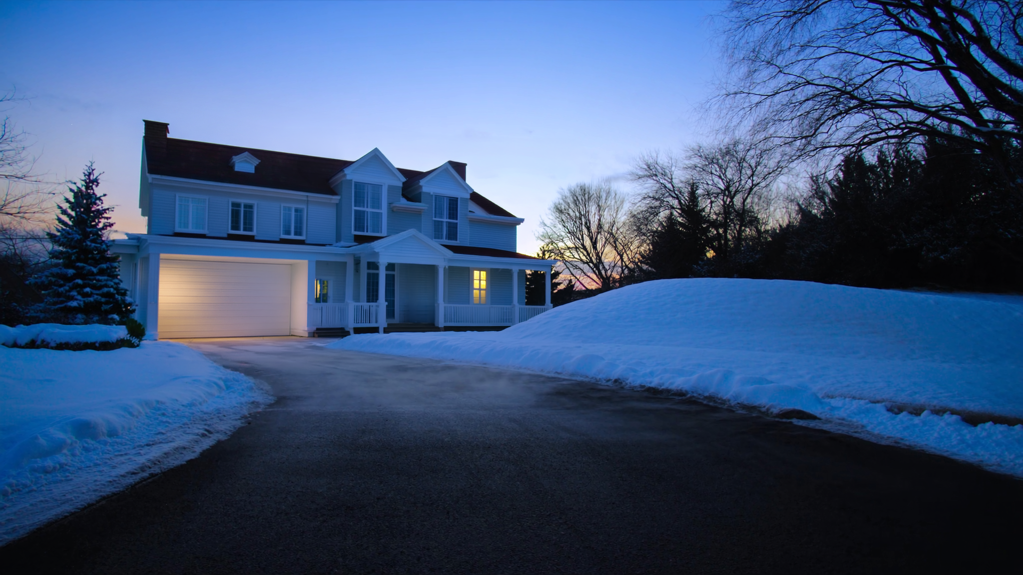 A residential driveway perfectly cleared of snow at blue-hour first light, leading up toward a white colonial home with the porch light glowing, plowed snow banks on both sides and mature trees flanking the property.